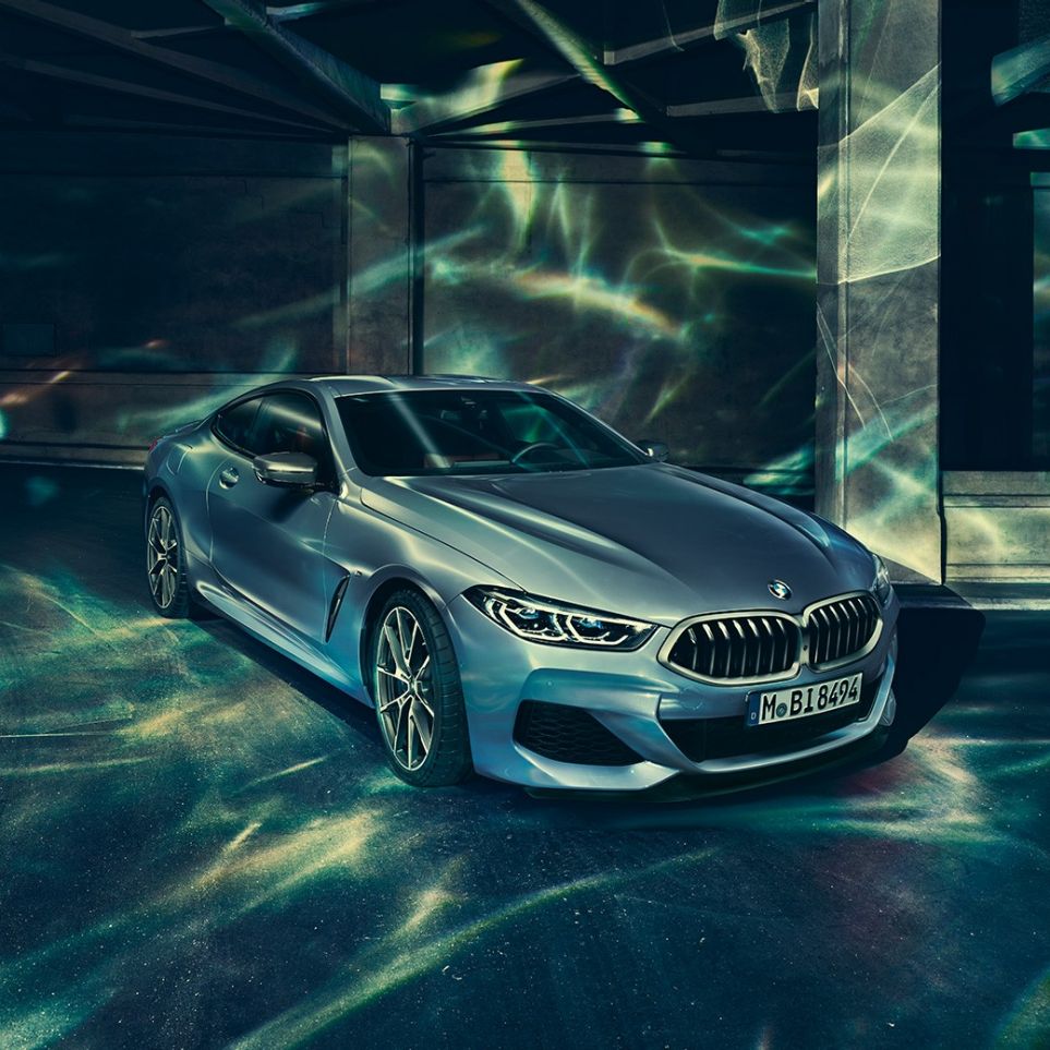 BMW M850i xDrive Coupé, вид спереди с поворотом в три четверти слева. BMW M850i xDrive Coupé в Barcelona Blue металлик, вид спереди с поворотом в три четверти слева, стоит перед зданием.