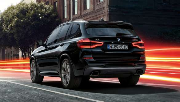 BMW X3 M40i, вид сзади с поворотом в три четверти. BMW X3 M40i в Black Sapphire металлик, за рулем, экстерьер, вид сзади с поворотом в три четверти на городском фоне со световыми полосками.