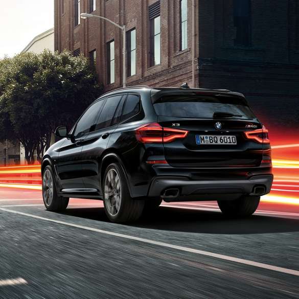 BMW X3 M40i, вид сзади с поворотом в три четверти. BMW X3 M40i в Black Sapphire металлик, за рулем, экстерьер, вид сзади с поворотом в три четверти на городском фоне со световыми полосками.