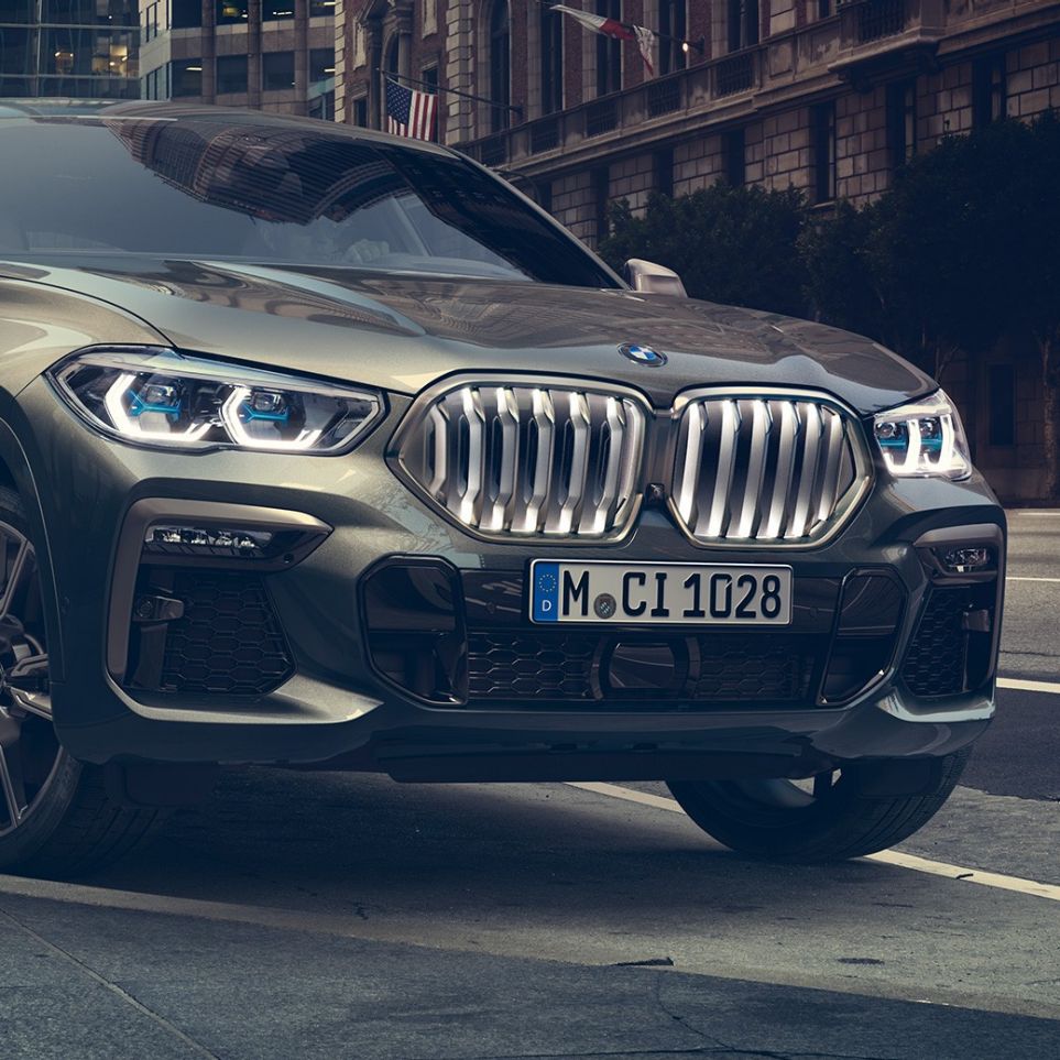 Неподвижно стоящий BMW X6 M50i BMW X6 M50i: вид спереди с поворотом в три четверти на фоне городского пейзажа
