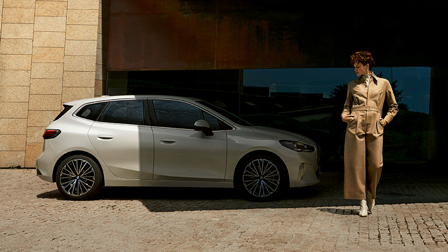 BMW 2 серии Active Tourer U06 2021 вид сбоку с моделью-женщиной BMW 2 серии Active Tourer U06 2021 в цвете Mineral White металлик на парковке, вид сбоку с моделью-женщиной