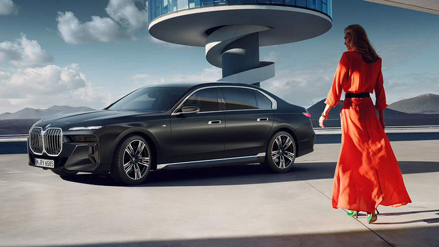 Модель в красном платье на фоне BMW 7 серии G70 ICE с пакетом M Sport BMW 7-series G70 ICE Пакет M Sport, Frozen Deep gray metallic, 21 легкосплавные диски M с двойными спицами 908, вид три четверти спереди с моделью в красном платье