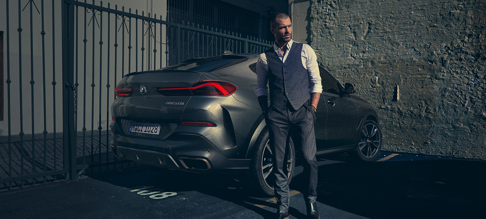 BMW X6 M50i, вид сзади с поворотом в три четверти. Мужчина стоит рядом с BMW X6 M50i SUV, вид сзади с поворотом в три четверти
